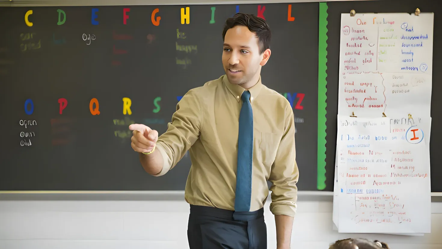 A man standing in front of a blackboard in a classroom. He is wearing a shirt, tie, and pants, and there are papers attached to the wall behind him. On the right side of the image, there is a board with something written on it.