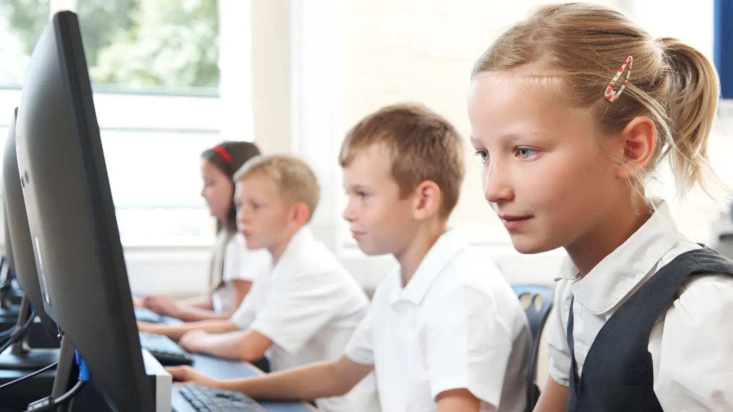 A group of children sitting at desks in front of a computer, with monitors, keyboards, mouses, and other objects on the tables. In the background, there is a wall with a window, through which trees can be seen.