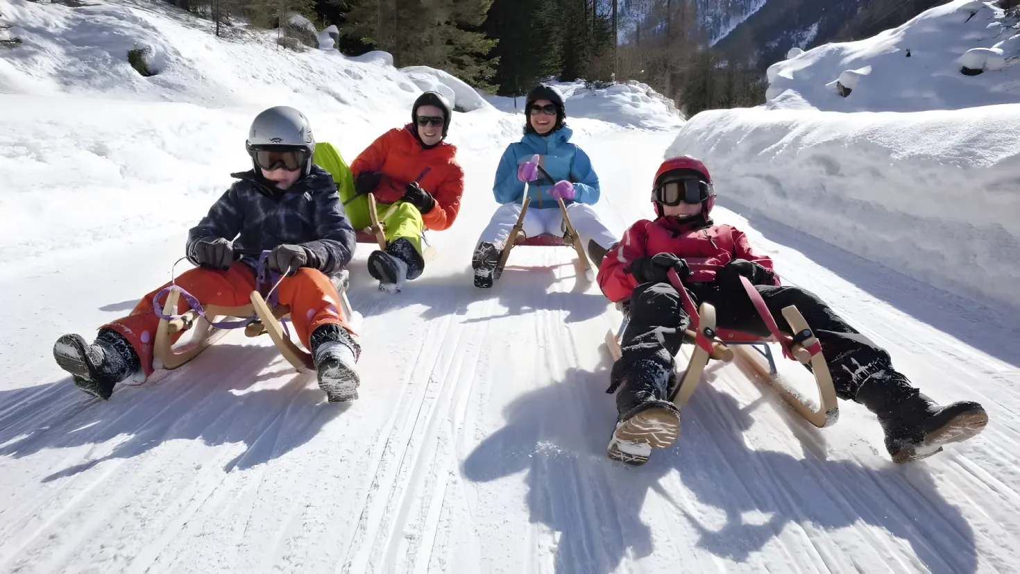 A group of children sledding down a snowy hill, wearing helmets, gloves, and sneakers. The background of the image is filled with trees and hills covered in snow, and the sky is visible at the top.